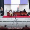 A woman in a headscarf speaks from a glass podium on a stage as a panel sits behind a long desk, Libyan flag nearby, and an audience watches.