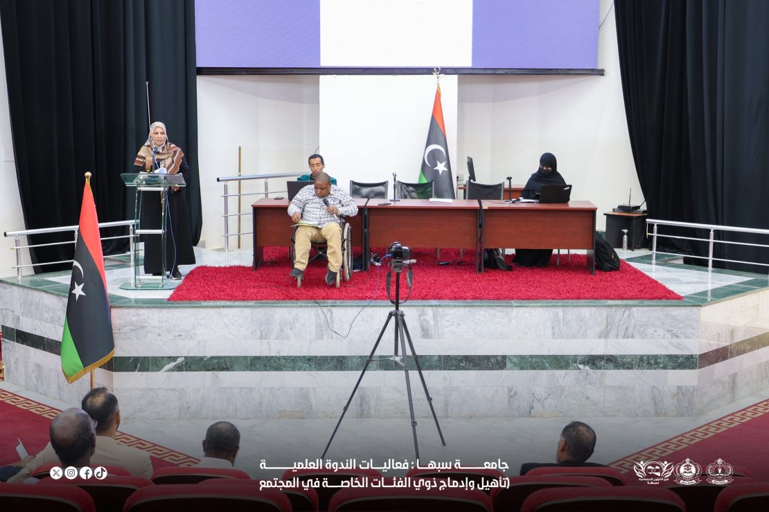 A woman in a headscarf speaks from a glass podium on a stage as a panel sits behind a long desk, Libyan flag nearby, and an audience watches.