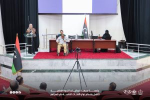 IMG-20261029-WA0008 - جامعة سبها | Sebha University A woman in a headscarf speaks from a glass podium on a stage as a panel sits behind a long desk, Libyan flag nearby, and an audience watches.