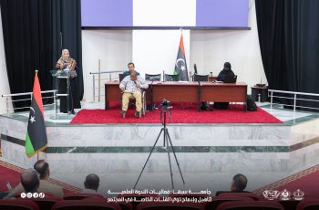 A woman in a headscarf speaks from a glass podium on a stage as a panel sits behind a long desk, Libyan flag nearby, and an audience watches.