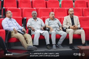 IMG-20261029-WA0010 - جامعة سبها | Sebha University Four men sit in red theater seats at a formal event, wearing business casual clothing and looking ahead.