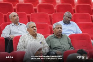 IMG-20261029-WA0011 - جامعة سبها | Sebha University Four men sit in red theater seats watching an event, looking toward the stage with attentive expressions, the foreground partially obscured by a caption overlay in Arabic at the bottom.