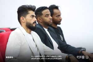 IMG-20261029-WA0012 - جامعة سبها | Sebha University ثلاثة رجال يجلسون في صف خلال فعالية رسمية، الأول في الأمام ببدلة بيضاء والثاني والثالث ببدلات داكنة يركزون على شيء أمامهم.