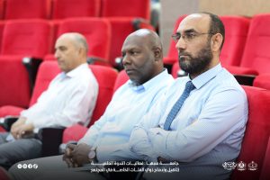 IMG-20261029-WA0013 - جامعة سبها | Sebha University Three men in business attire seated in red auditorium seats, attentively watching a presentation.