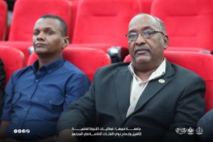 IMG-20261029-WA0015 - جامعة سبها | Sebha University Two men sit in red auditorium chairs at a formal event, one in a blue shirt and the other in a blazer with glasses, foreground and background visible.