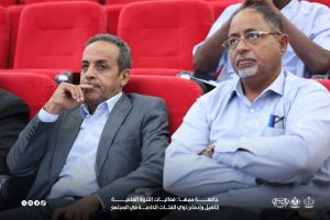 IMG-20261029-WA0024 - جامعة سبها | Sebha University Two men in business attire seated in red auditorium chairs, attentively watching an event.