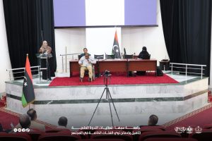 IMG-20261029-WA0025 - جامعة سبها | Sebha University Panel discussion in a hall with Libyan flags, a speaker at a podium on the left, a man in a wheelchair seated at the center table, and a woman in hijab at a laptop on the right during a formal event.
