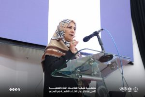 IMG-20261029-WA0026 - جامعة سبها | Sebha University Woman wearing a patterned hijab speaks at a glass podium with a microphone during a formal event.