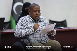 IMG-20261029-WA0029 - جامعة سبها | Sebha University Man in a wheelchair speaks into a handheld microphone while reading from a sheet of paper at a conference or meeting room.