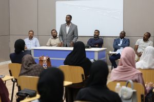 IMG_0530 - جامعة سبها | Sebha University Man in a gray plaid blazer standing at a front desk, addressing a panel of five men in a classroom setting.