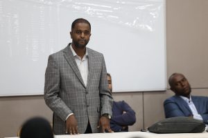 IMG_0532 - جامعة سبها | Sebha University Man in a gray plaid suit stands at a conference table, presenting or speaking, with colleagues seated in the background in a meeting room.