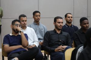 IMG_0539 - جامعة سبها | Sebha University Group of men seated in a panel or classroom, facing forward and listening intently in a neutral gray-room setting.