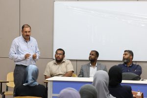 IMG_0550 - جامعة سبها | Sebha University A man in a checkered shirt stands at the front of a classroom addressing seated students in hijabs and casual outfits, with a whiteboard behind him.