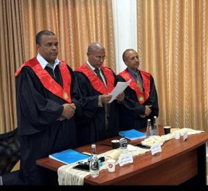 Three men in black academic gowns with red stoles stand behind a table, one reading a document as others listen in a formal ceremony.