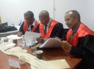 Three judges in black robes with red sashes review documents at a table, with papers, water bottles, and coffee cups on the surface.