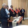 Group of men in a formal ceremony around a table with gift bags and cups; three wear red-and-black academic gowns, others in suits.