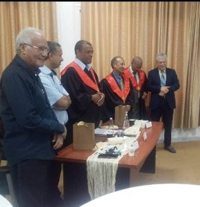 WhatsApp Image 2026-10-04 at 11.07.04 PM - جامعة سبها | Sebha University Group of men in a formal ceremony around a table with gift bags and cups; three wear red-and-black academic gowns, others in suits.