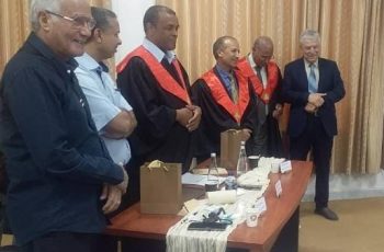 Group of men in a formal ceremony around a table with gift bags and cups; three wear red-and-black academic gowns, others in suits.