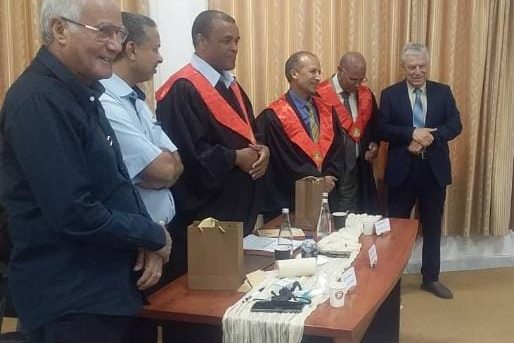 Group of men in a formal ceremony around a table with gift bags and cups; three wear red-and-black academic gowns, others in suits.