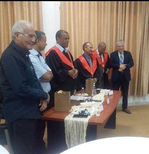 WhatsApp Image 2026-10-04 at 11.07.04 PM - جامعة سبها | Sebha University Group of men in a formal ceremony around a table with gift bags and cups; three wear red-and-black academic gowns, others in suits.