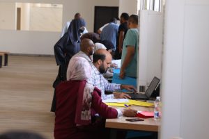 People seated at a long table with laptops and forms, assisting individuals in a registration line across a spacious room?