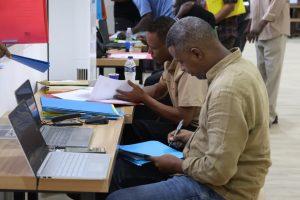 Two men sit at a registration desk with laptops, folders, and papers, filling out forms.