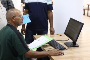 Man seated at a desk using a desktop computer, with papers in hand and two people standing nearby.