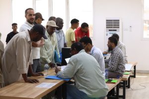Group of men line up to register at a desk where staff sign documents in a bright room.