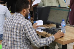 Man in a plaid shirt sits at a desk, holding a receipt while using a desktop computer; a water bottle and keyboard are on the desk.