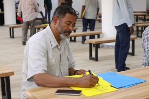Man with sunglasses on his head writes on bright yellow sheets at a wooden table in a large, open space with benches and other people in the background.