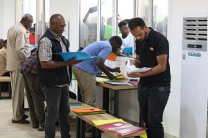 Men stand at a service desk sorting colorful folders and papers in a bright room as others wait their turn