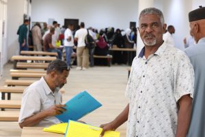 Smiling man in a patterned shirt stands in a bright hall while another man sits at a bench reading blue and yellow folders, with a long line of people in the background.