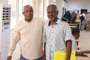 Two smiling men stand side by side indoors, one with his arm around the other's shoulder.