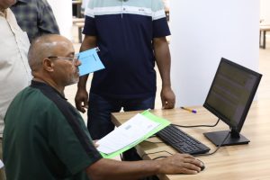 Older man with glasses sits at a desk using a desktop computer while two coworkers stand nearby with documents in hand.