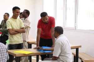 Man in a red shirt leans over a desk to talk with a seated man while others wait in a bright room with windows in the background.