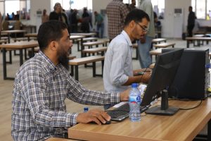 Two men sit at a long wooden table using laptops and a monitor in a busy open workspace; a water bottle sits between them.