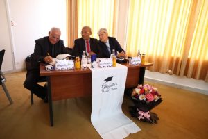 WhatsApp Image 2026-10-04 at 7.43.33 PM (1) - جامعة سبها | Sebha University Three men in suits signing documents at a wooden table during a formal thesis event, with a white banner reading'Master's Thesis' draped over the edge and a bouquet of flowers nearby on the floor to the right.