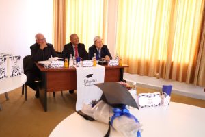 WhatsApp Image 2026-10-04 at 7.43.36 PM - جامعة سبها | Sebha University Three men in suits sit at a panel table during a formal event, with a graduation-themed banner and gifts on the table in a sunlit room.