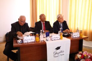 WhatsApp Image 2026-10-04 at 7.43.38 PM (2) - جامعة سبها | Sebha University Three men in suits sit at a conference table during a master's thesis defense, with name cards and orange juice bottles in front of them.
