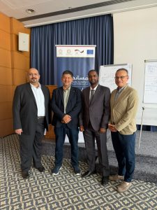 Four men in business attire stand together posing for a photo at an indoor event, with a blue banner and whiteboards in the background.