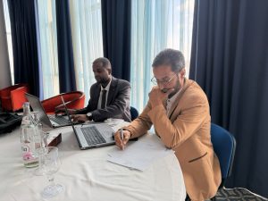 Two men in business attire work at a round table with laptops and documents in a bright room with blue curtains.