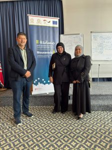 Three professionals pose for a photo beside a Mousanada II banner at a conference, with whiteboards in the background.