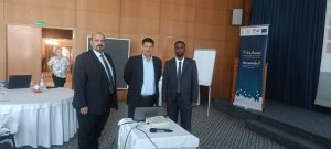 Three men in suits pose for a photo in a conference room, with a projector and Mousanad II banner in the background.