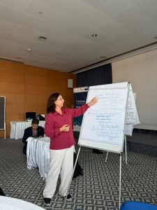 Woman in a pink blouse standing beside a white flip chart, presenting to a conference room with a seated man in the background.