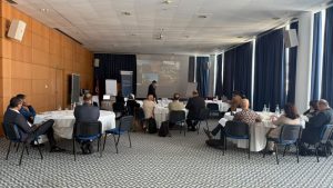 Conference room with round tables and attendees listening to a presenter at the front, projection screen visible ahead.
