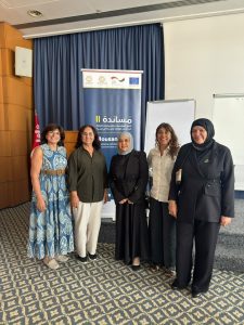 WhatsApp Image 2026-10-04 at 8.34.15 PM - جامعة سبها | Sebha University Five women pose for a photo in a conference room, standing in front of a blue-curtained stage with a banner that has logos and Arabic text behind them.
