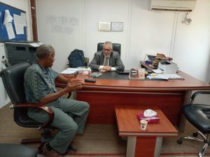 Two men talk across a large office desk, with papers and a printer nearby, in a small meeting room.
