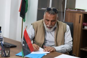 Man with a gray beard signs papers at a desk in an office; a red-black-green flag on the desk and sunglasses atop his head.