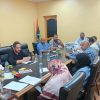 A business meeting around a long conference table with ten people in an office, several water bottles, papers, and a Libyan flag in the background.