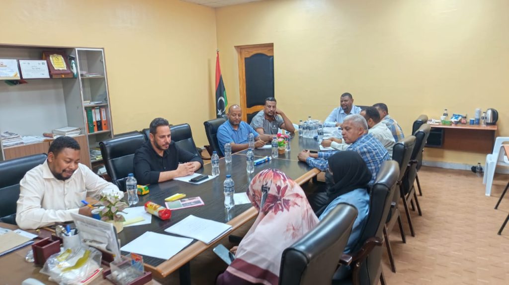 WhatsApp Image 2026-10-13 at 12.07.46 PM (2) - جامعة سبها | Sebha University A business meeting around a long conference table with ten people in an office, several water bottles, papers, and a Libyan flag in the background.